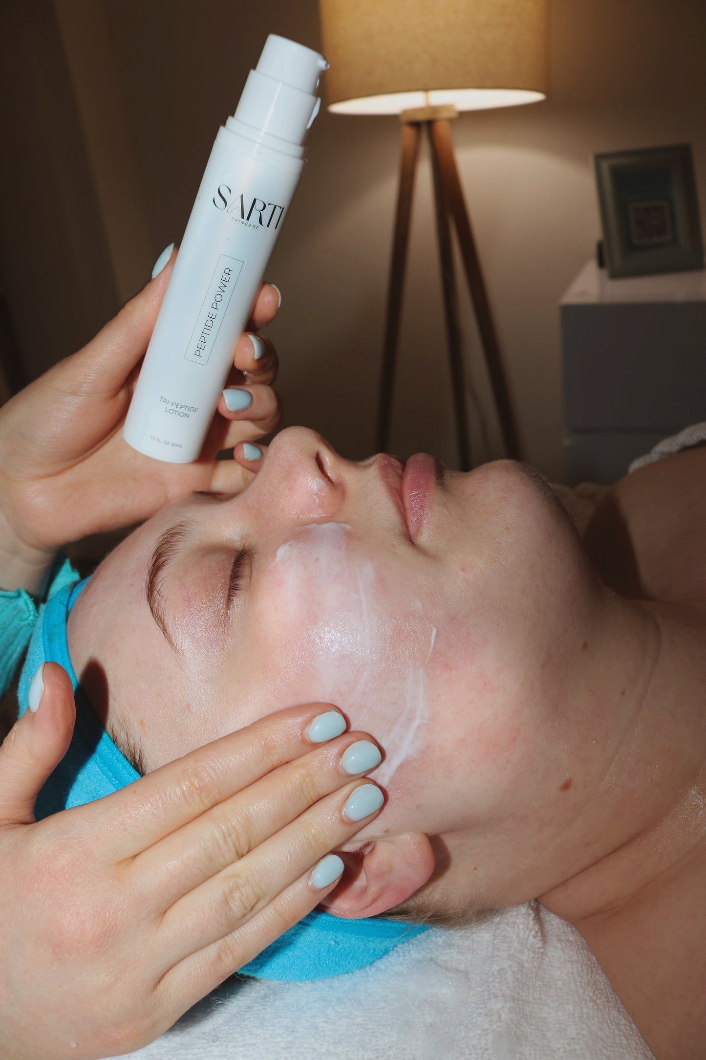 Person applying a facial product to another person's face with a lamp in the background